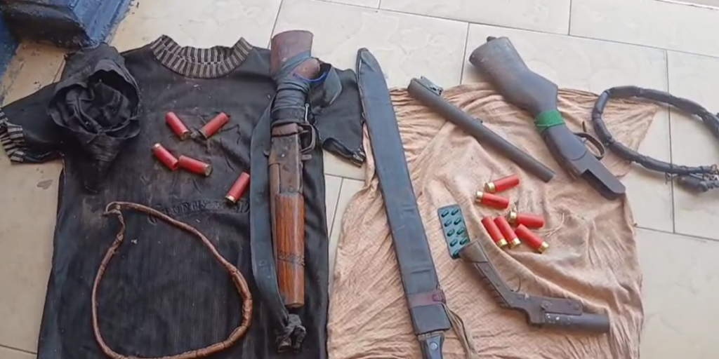 image-1013-1024x512 | Ghanaguardian.com Collection of weapons and ammunition laid out on cloth: shotgun, magazines, red shotgun shells, and a sheathed blade on a tiled floor, likely for display or inspection.