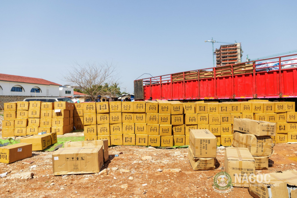 Stacked cardboard boxes at a construction site beside a red trailer truck under a blue sky.