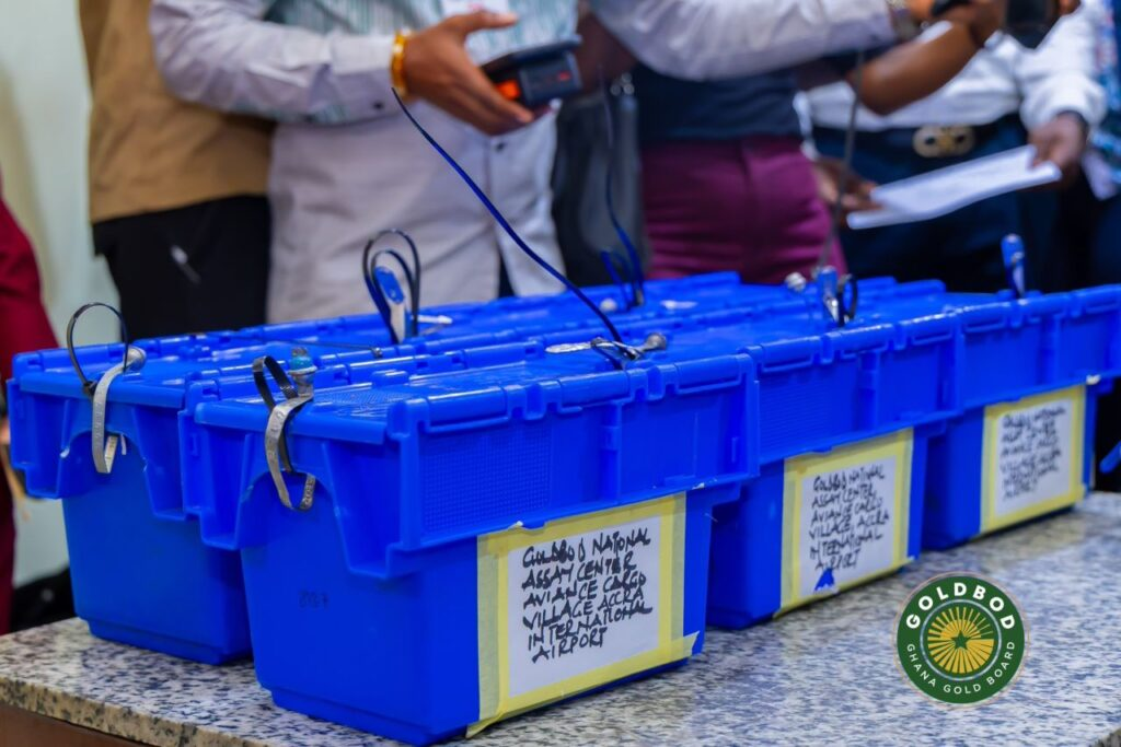 Row of blue plastic storage boxes with labeled tapes on a speckled counter; people stand behind, handling a device. A circular Gold Board Ghana logo is visible on the lower right.