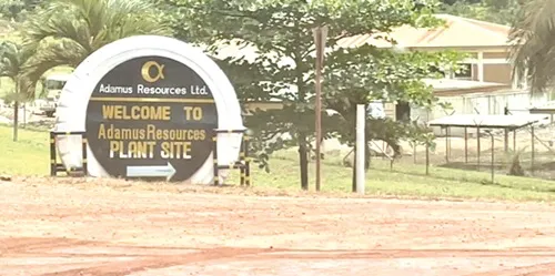 Circular sign reading 'Welcome to Adamus Resources Plant Site' with arrow, set on a dirt road near trees and a building in the background.