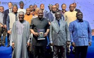 Group of diverse officials posing on a stage; central man in black shirt holds a booklet in front of a blue backdrop with other attendees around him.