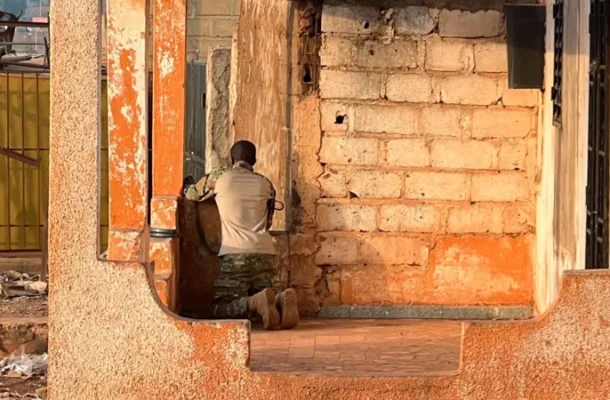 Man kneels with back to camera beside a weathered brick wall inside a ruined building; wearing light shirt and camouflage pants.