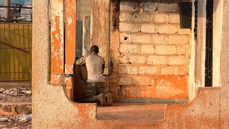Man kneels with back to camera beside a weathered brick wall inside a ruined building; wearing light shirt and camouflage pants.