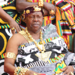 Elderly African king seated, adorned in gold jewelry and a patterned robe, with attendants in colorful fabrics nearby.