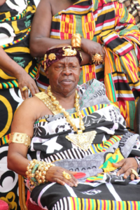 Elderly African king seated, adorned in gold jewelry and a patterned robe, with attendants in colorful fabrics nearby.