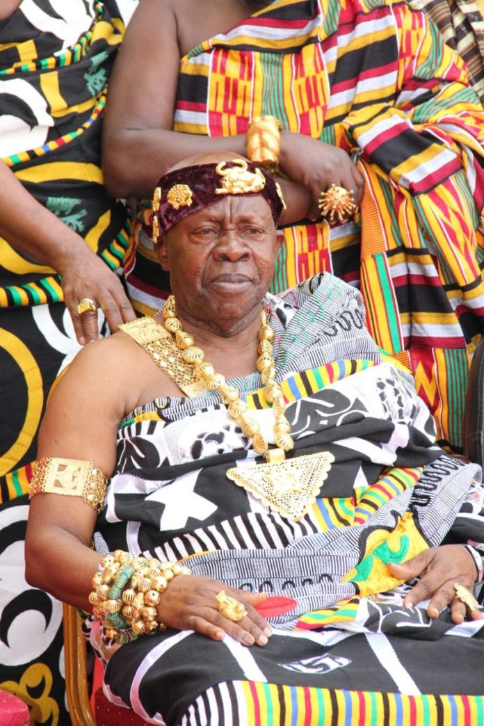 Elderly African king seated, adorned in gold jewelry and a patterned robe, with attendants in colorful fabrics nearby.