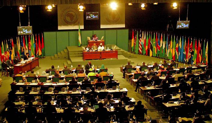 Large international conference hall with delegates seated at long desks, numerous national flags lining the walls behind the stage.