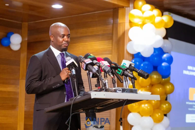 Man in a dark suit speaking at a podium with multiple microphones during a press conference, balloons in gold, white, and blue in the background