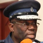 Police officer in a dark blue uniform and peaked cap speaks into orange and blue microphones at a press event.