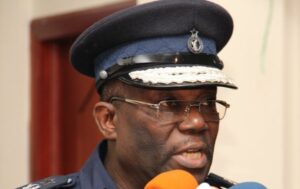 Police officer in a dark blue uniform and peaked cap speaks into orange and blue microphones at a press event.