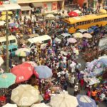 Crowded urban market with dense stalls under colorful umbrellas, shoppers, vendors, and two yellow buses on a busy street.