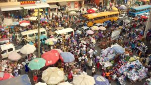 Crowded urban market with dense stalls under colorful umbrellas, shoppers, vendors, and two yellow buses on a busy street.