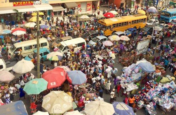 Crowded urban market with dense stalls under colorful umbrellas, shoppers, vendors, and two yellow buses on a busy street.