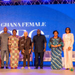 Nine people stand on a stage in front of a blue screen that reads 'GHANA FEMALE', wearing traditional and formal outfits.