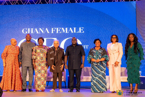 Nine people stand on a stage in front of a blue screen that reads 'GHANA FEMALE', wearing traditional and formal outfits.