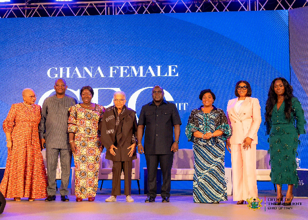 Nine people stand on a stage in front of a blue screen that reads 'GHANA FEMALE', wearing traditional and formal outfits.