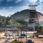 Industrial mining site featuring a tall headframe, low buildings, and several trucks parked near dirt roads with hillside backdrop