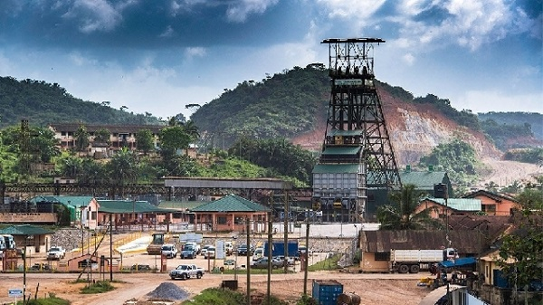 Industrial mining site featuring a tall headframe, low buildings, and several trucks parked near dirt roads with hillside backdrop