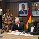 Two men sign documents at a table while four colleagues stand behind, in a room with Ghanaian flags and portraits on the wall.