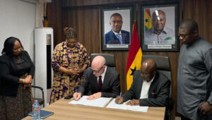 Two men sign documents at a table while four colleagues stand behind, in a room with Ghanaian flags and portraits on the wall.