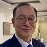 Close-up of a smiling man wearing glasses and a dark suit at an indoor event, with framed artwork on the wall in the background.