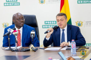 Two men in suits at a press briefing table, microphones in front; one speaks into a handheld mic, flag visible in the background.