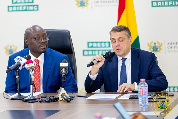 Two men in suits at a press briefing table, microphones in front; one speaks into a handheld mic, flag visible in the background.