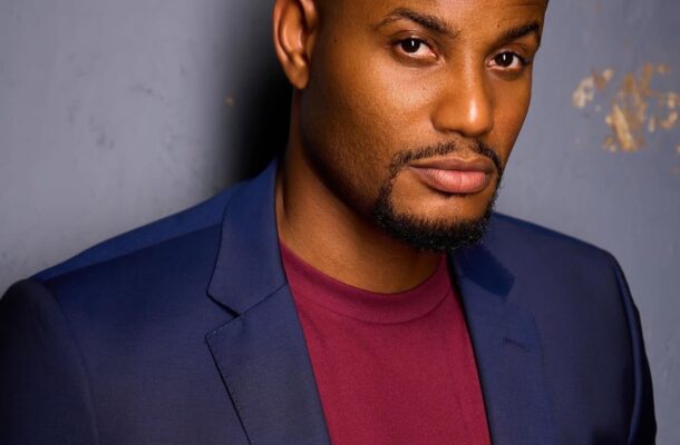Portrait of a man wearing a navy blazer and maroon shirt, posing against a gray wall.