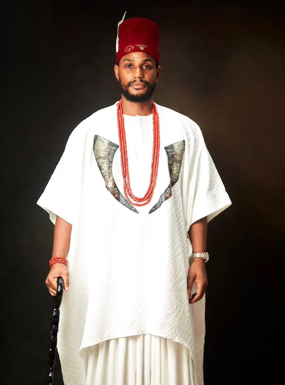 Man in a white agbada with a red hat (fila), red beaded necklaces, a red bracelet, and a silver watch, standing against a dark background while holding a black cane.