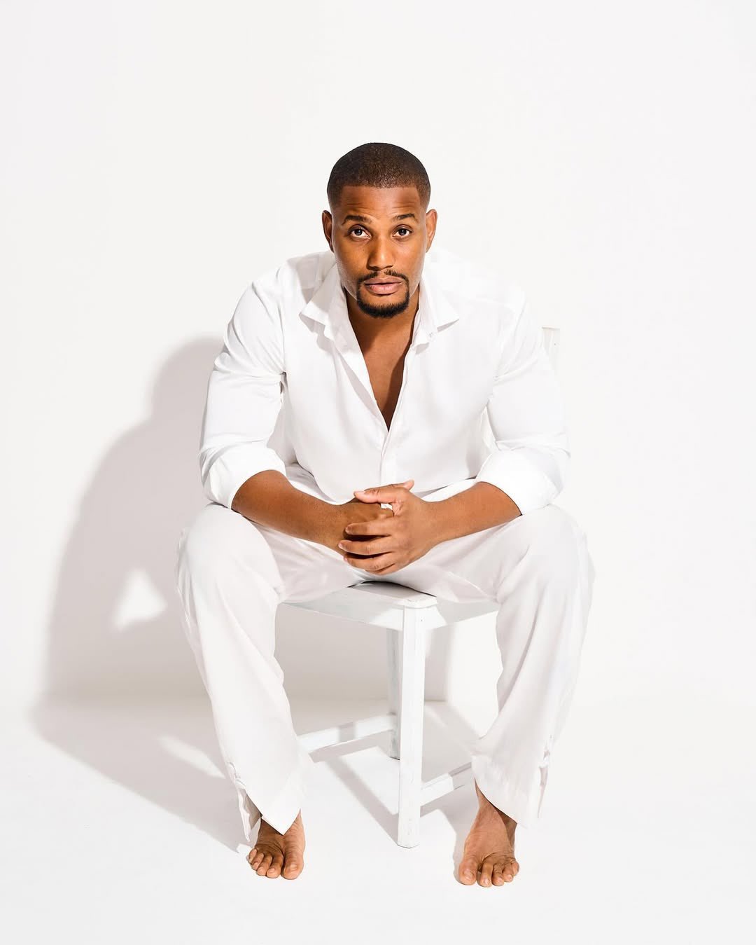 Portrait of a man in a white shirt and pants sitting barefoot on a white stool in a bright studio, hands clasped and looking at the camera.