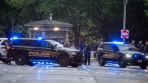 Two police SUVs with flashing blue lights block a street with bystanders and investigators in the background near a small guardhouse park entrance.