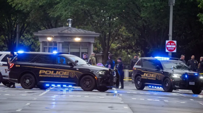 Two police SUVs with flashing blue lights block a street with bystanders and investigators in the background near a small guardhouse park entrance.