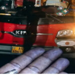 Close-up of a gloved hand with a ring in front of a red Kia car with headlights on at night, license plate AS 4941 visible.