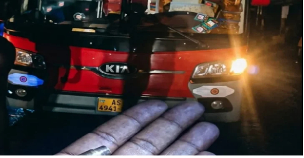 Close-up of a gloved hand with a ring in front of a red Kia car with headlights on at night, license plate AS 4941 visible.