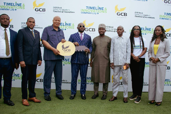 Group of eight people in business attire posing on a green carpet at a corporate event, with a GCB circular sign and a timeline innovations backdrop.