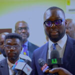Man in a dark suit and glasses speaks into microphones at a press conference, with reporters and others in the background.