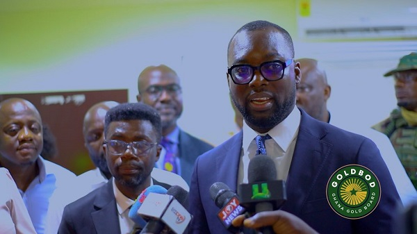 Man in a dark suit and glasses speaks into microphones at a press conference, with reporters and others in the background.