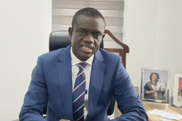 Professional man in a blue suit and striped tie sits at an office desk, looking at the camera with framed photos on the desk behind him.