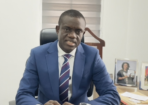 Professional man in a blue suit and striped tie sits at an office desk, looking at the camera with framed photos on the desk behind him.