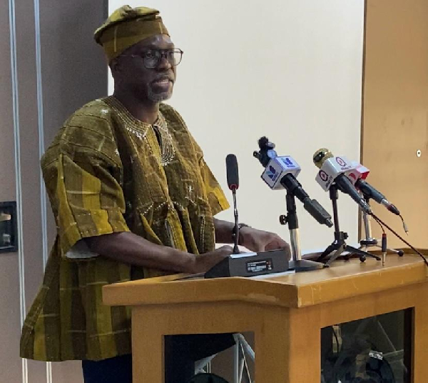 Man in traditional African attire speaks at a wooden podium with several microphones in front of him at a press conference/Flex event hall.
