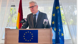 Man in a suit and glasses speaks at a lectern with the EU flag emblem, microphones in front, flanked by a German flag and an EU flag behind him.