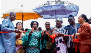 Diverse group of people holding a red ribbon together at an outdoor ribbon-cutting ceremony under colorful umbrellas.