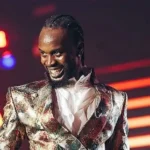 Male performer on stage wearing a shiny metallic jacket, smiling as neon lights glow in the background.