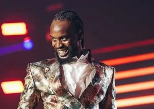 Male performer on stage wearing a shiny metallic jacket, smiling as neon lights glow in the background.