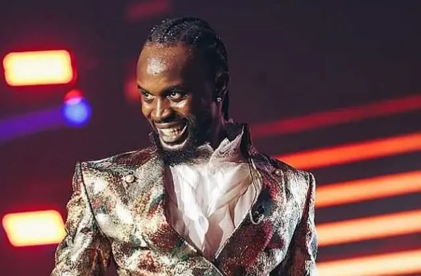 Male performer on stage wearing a shiny metallic jacket, smiling as neon lights glow in the background.