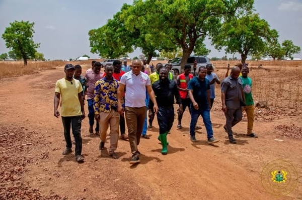 Group of men walking toward the camera along a dusty red dirt road in a rural landscape, with trees and a vehicle in the background.