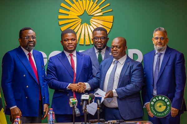 Five men in suits at a press conference; two central figures shake hands and present documents, microphones in front, green backdrop with a sunburst emblem.