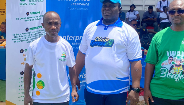 Three men posing at an outdoor promotional booth; left in a white T-shirt with logos, center in a white-blue polo, right in a green shirt, with banners and seated spectators behind.