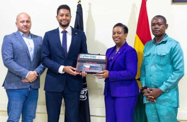Four adults in business attire pose indoors as a boxed CyberVAC device is handed over during a formal ceremony. The flag-topped background adds a official setting.