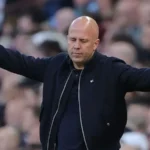 Bald man in a black jacket stands with arms spread wide on a sports field, eyes closed amid a cheering crowd behind him.